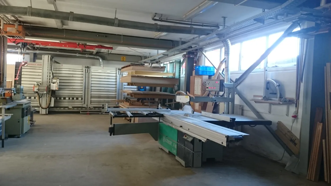 Blick in den Maschinenpark: Formatkreissäge im Vordergrund, dahinter Plattensäge, Absaugung und Regale mit Plattenmaterial