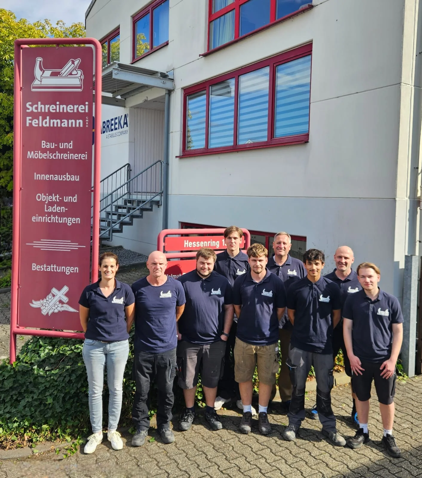 Das Team der Schreinerei Feldmann vor dem Betrieb am Hessenring: neun Kolleginnen und Kollegen in dunkelblauen Firmenhemden neben dem roten Firmenschild mit dem Hobel